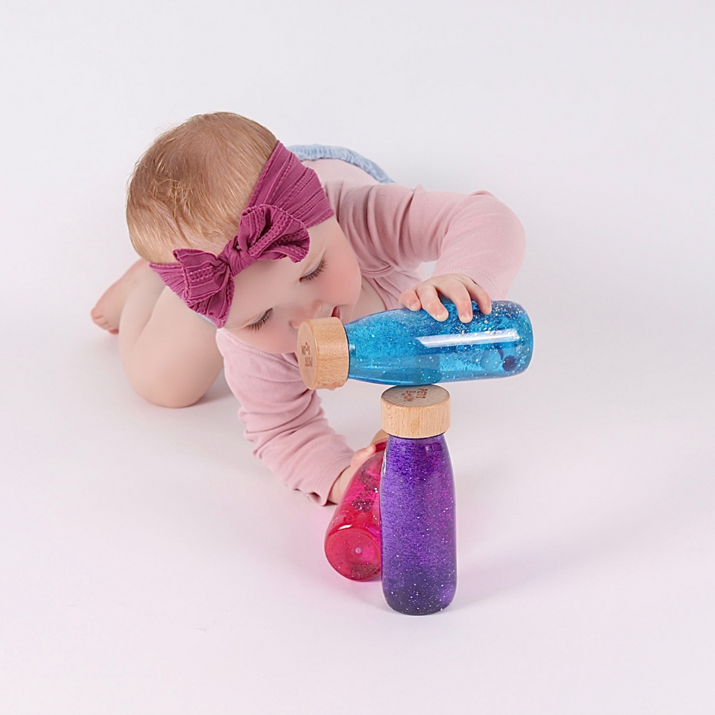 Baby playing with the Petit Boum Magic Pack which includes a Petit Boum pink sensory float bottle, a purple sensory float bottle and a blue sensory float bottle.