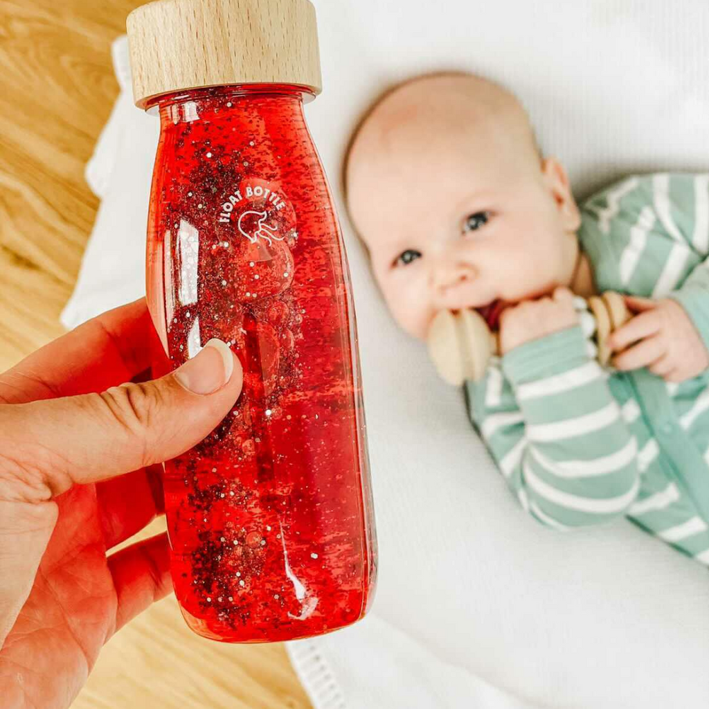 A baby holding a wooden beaded teething rattle whilst being shown a red Petit Boum sensory bottle. The toys are from the 3-6 month baby's first year subscription box.