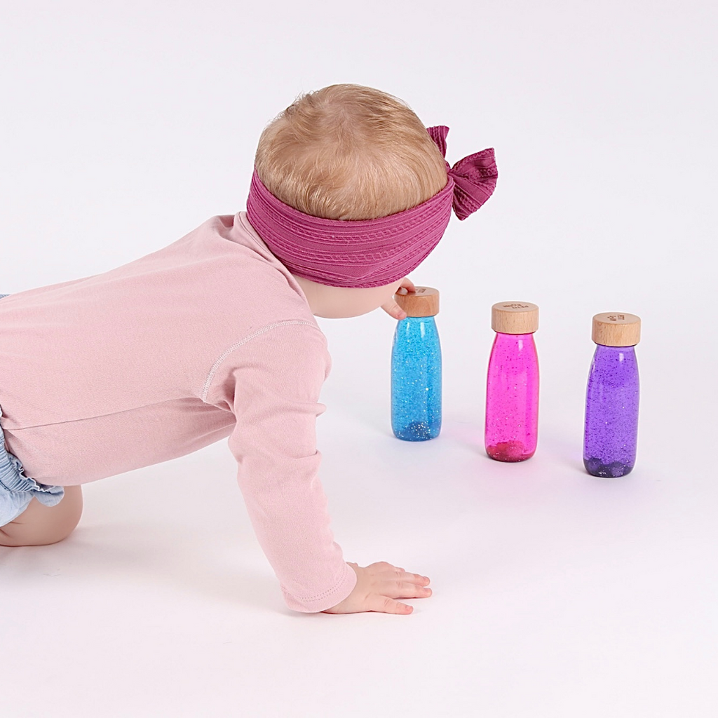 Baby playing with the Petit Boum Magic Pack which includes a Petit Boum pink sensory float bottle, a purple sensory float bottle and a blue sensory float bottle. All bottles lined up next to each other.