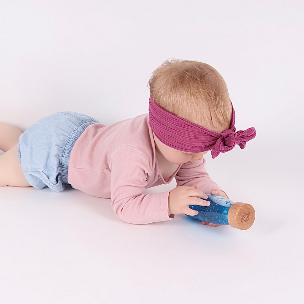 Baby playing with the Petit Boum Magic Pack during tummy time which includes a Petit Boum pink sensory float bottle, a purple sensory float bottle and a blue sensory float bottle.