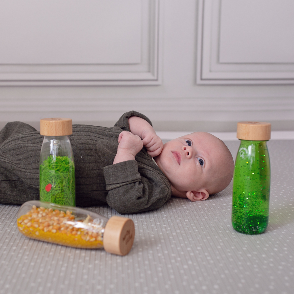 A baby laying on it's back looking at the Petit Boum life pack which includes a green sensory float bottle, a farm spy sensory bottle and a ladybird sound sensory bottle.