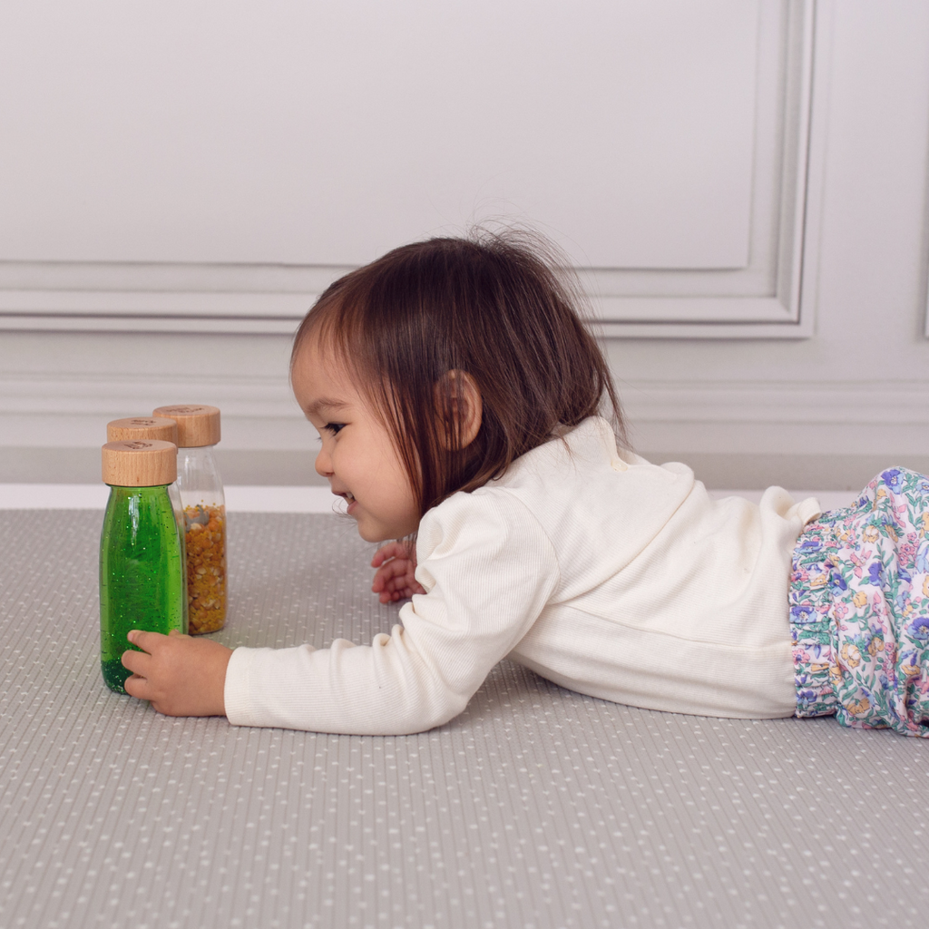A toddler laying on their front and holding the Petit Boum life pack sensory bottles.