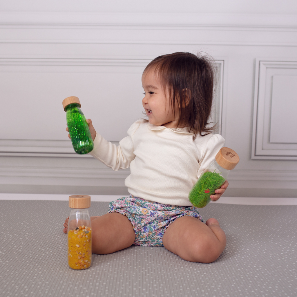 A toddler knealing and holding a green Petit Boum sensory float bottle and a ladybird sound sensory bottle.