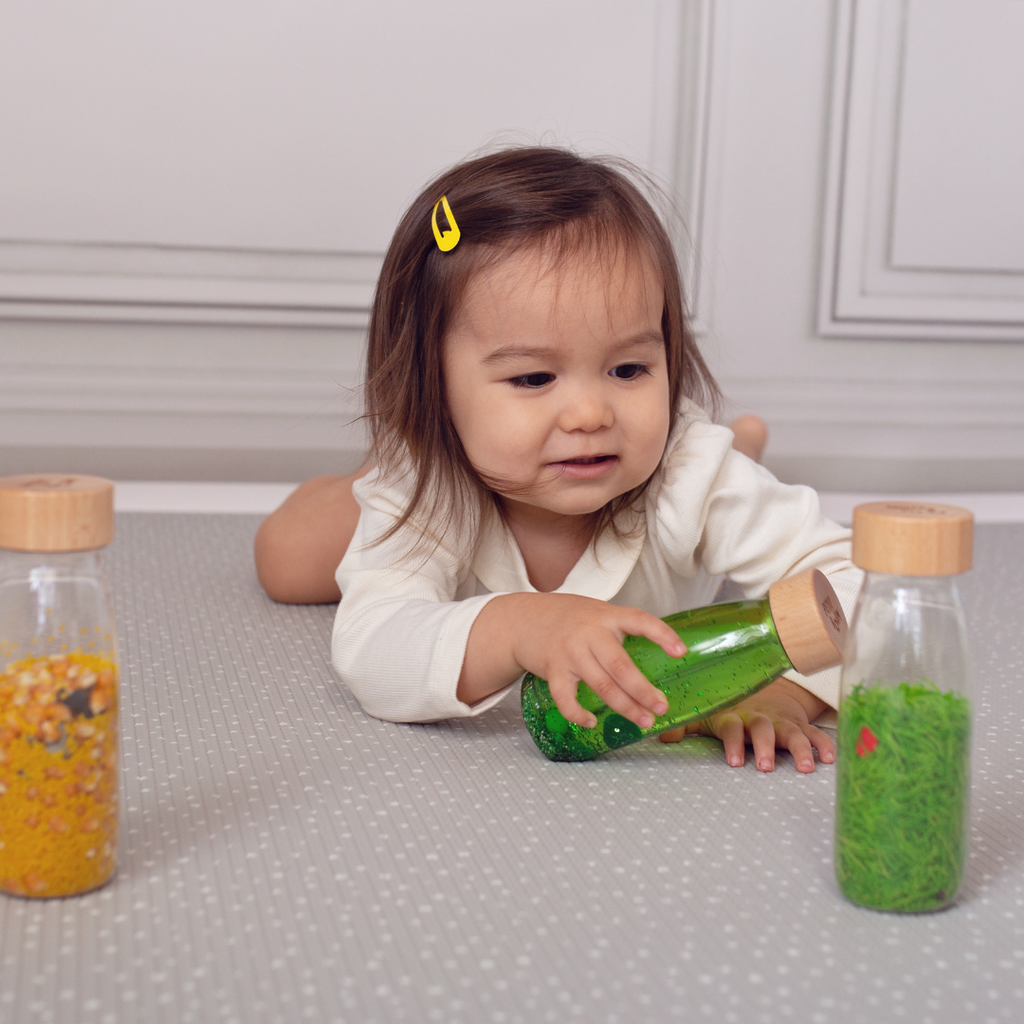 A toddler laying on their front looking and playing with the Petit Boum life pack which includes a green sensory float bottle, a farm spy sensory bottle and a ladybird sound sensory bottle.